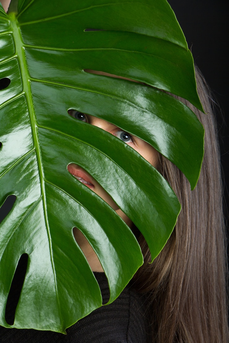 Foto hermosa chica morena con cabello largo mira a través de hojas de monstera en negro