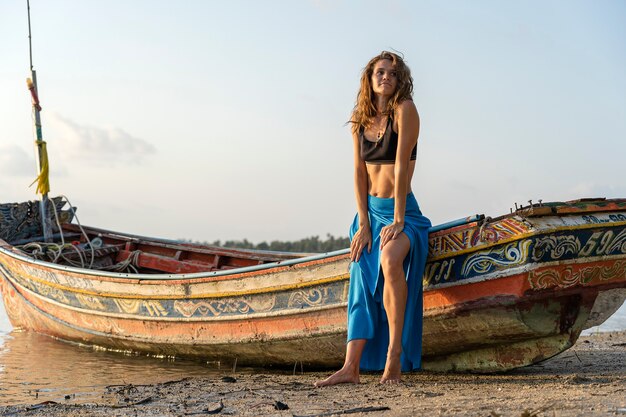 Hermosa chica morena caucásica en la playa de arena tropical cerca de barco de madera al atardecer, Tailandia, de cerca. Concepto de naturaleza y verano.