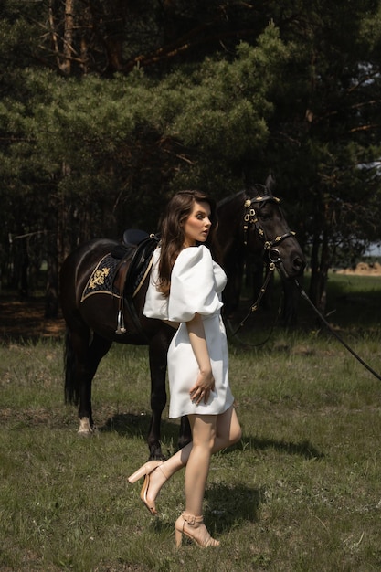 Hermosa chica morena caucásica con un vestido de novia se encuentra junto a un caballo en el bosque