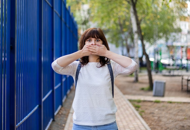 Una hermosa chica morena se cubrió la boca con las manos. no decir nada. gesto de silencio. una niña con una chaqueta blanca está de pie en la calle.