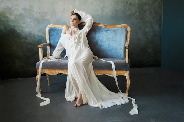 Hermosa chica morena delgada sentada en el sofá con un vestido blanco largo. Retrato de una mujer con una joya en el cuello. Peinado perfecto y cosmética de la mujer, nueva colección de vestidos ligeros