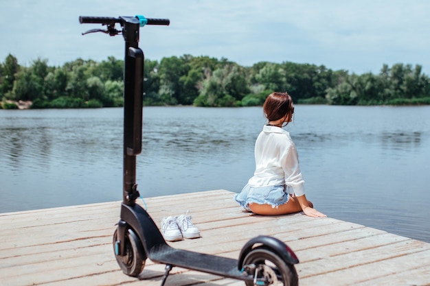 Una hermosa chica morena descansa sobre un puente de madera cerca del río junto a un scooter eléctrico. El concepto de ecología y transporte eléctrico.