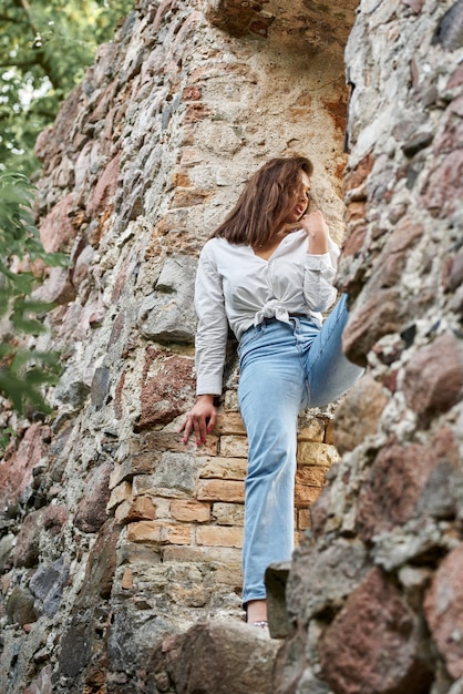 Hermosa chica morena en jeans y una camisa blanca posando sobre las ruinas del bosque