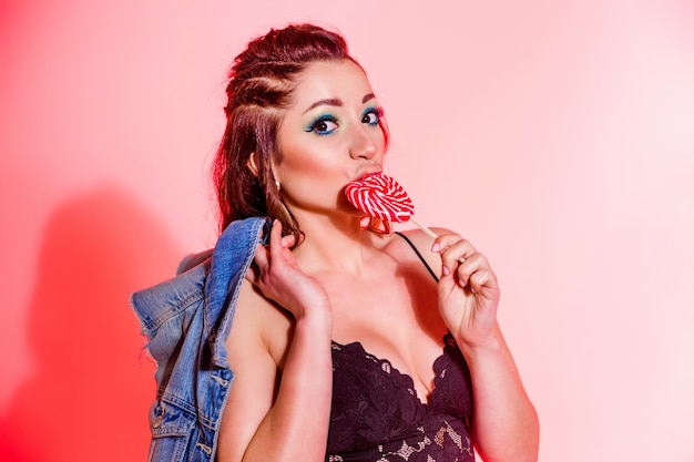 Hermosa chica morena con maquillaje azul, coletas y un top negro sexy posando sobre un fondo rojo con una piruleta en forma de corazón. Foto horizontal