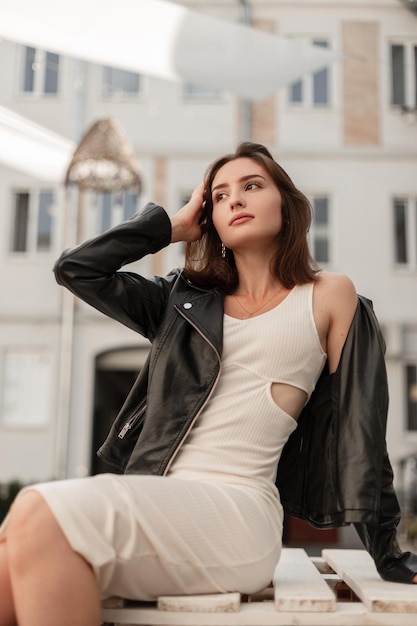 Hermosa chica morena de moda con un elegante vestido blanco con una chaqueta negra de cuero se sienta en paletas de madera en la ciudad