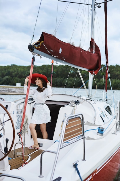 Hermosa chica morena modelo nd de moda en vestido corto y elegante blanco sonriendo, ajustando su sombrero rojo de moda y posando en un yate en el mar