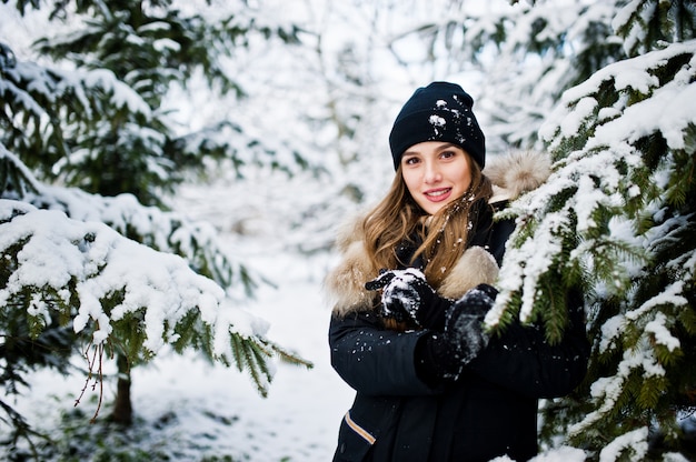 Hermosa chica morena en ropa de abrigo de invierno. Modelo con chaqueta de invierno y sombrero cerca de pinos.