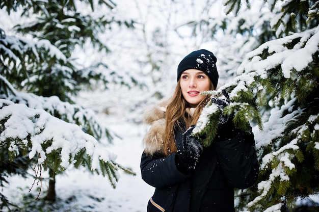 Hermosa chica morena en ropa de abrigo de invierno. Modelo en chaqueta de invierno y sombrero cerca de pinos.