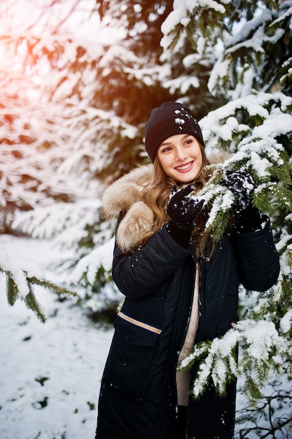 Hermosa chica morena en ropa de abrigo de invierno. Modelo en chaqueta de invierno y sombrero cerca de pinos.