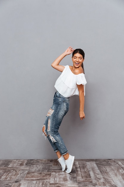 Hermosa chica morena sonriente alegre de pie de puntillas aislado en una pared gris