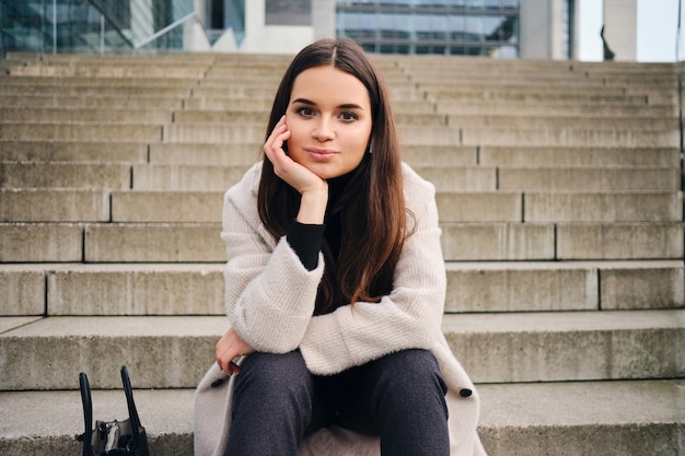 Hermosa chica morena sonriente mirando soñadoramente a la cámara descansando en las escaleras al aire libre