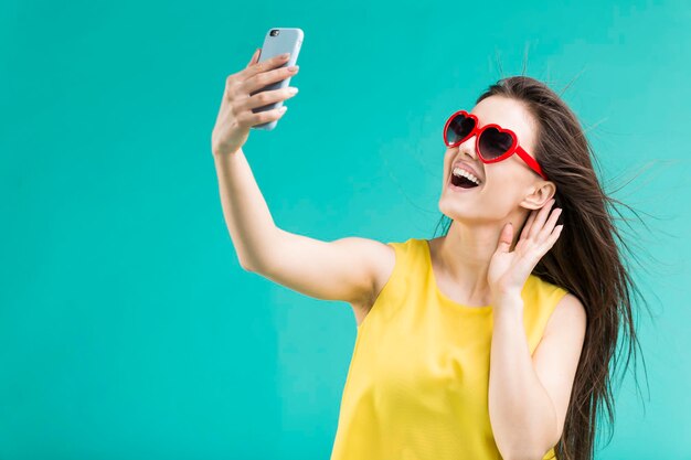 Hermosa chica morena sonriente usa camiseta amarilla y gafas de sol haciendo selfie por teléfono inteligente antes de fondo azul