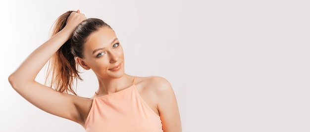 Foto una hermosa chica morena sostiene su cabello y hace su propio peinado. modelo joven con pelo largo, top naranja de moda sobre una superficie gris