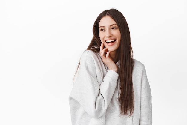 Hermosa chica morena tocándose la cara, riendo y mirando a un lado en el espacio de la copia del logo, mirando al lado izquierdo con expresión feliz y complacida, de pie sobre una pared blanca