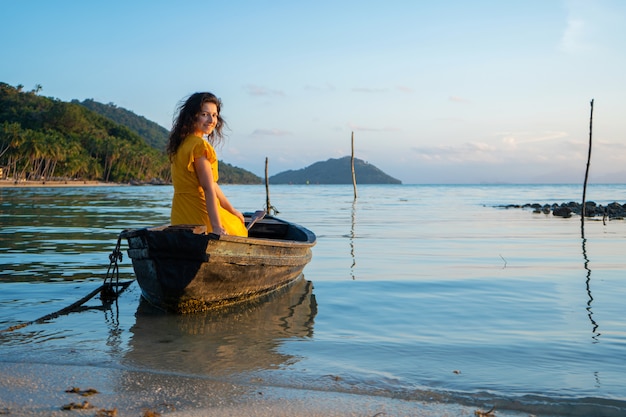 Hermosa chica morena con un vestido amarillo se sienta en un viejo bote de madera con vistas a una isla tropical desierta. Romance en el mar