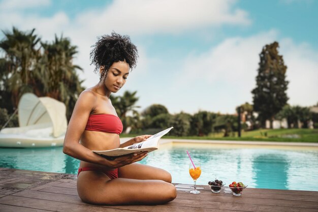 Una hermosa chica multirracial con cabello rizado en bicicleta roja relajándose sentada junto a la piscina leyendo una revista y tomando un desayuno saludable de frutas, cerezas y fresas, espacio para copiar