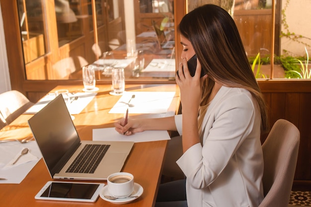 Hermosa chica de negocios hablando por teléfono mientras toma notas en un papel en el restaurante