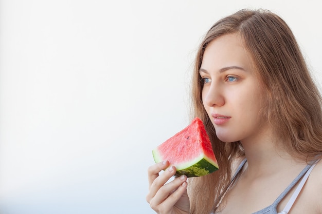 Hermosa chica de ojos azules con cabello largo castaño claro, en camiseta gris con tirantes, come sandía roja. Sobre fondo blanco. Copie el espacio.