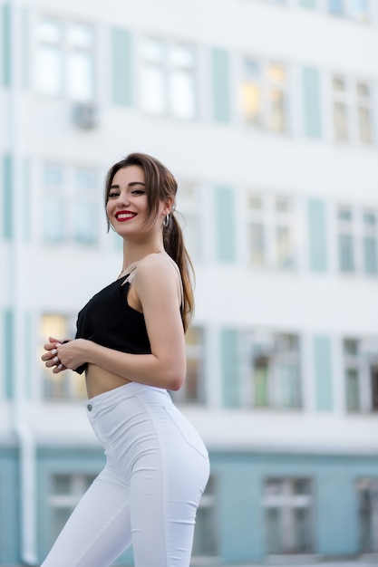 Una hermosa chica con pantalones blancos y un suéter negro posa con el telón de fondo de un edificio