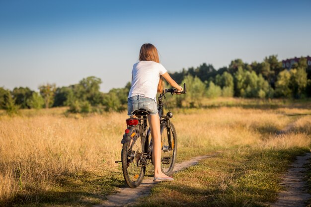 Hermosa chica en pantalones cortos y camiseta en bicicleta en la pradera al atardecer