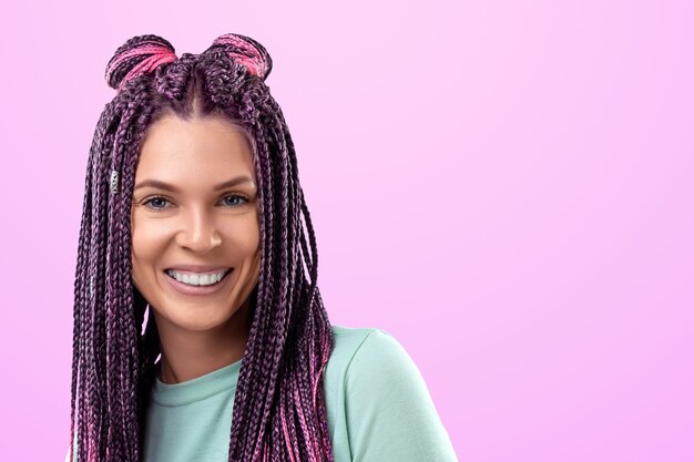 Hermosa chica con un peinado de trenzas rosas en ropa turquesa sonríe y posa sobre un fondo rosa en el estudio. El concepto es estilo moderno, individualidad, inclusividad, creatividad.