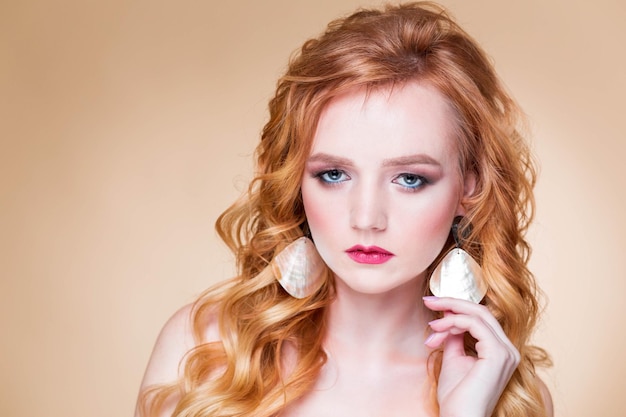 Una hermosa chica pelirroja con cabello ondulado y poses de maquillaje sobre un fondo beige en el estudio con aretes redondos de oro Mujer tocando el arete y mirando a la cámara