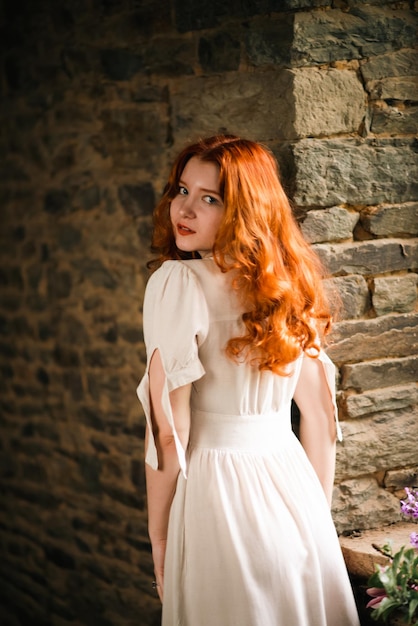 Hermosa chica pelirroja cerca de la ventana vieja con un vestido de noche blanco en una casa tradicional francesa el tema del romance