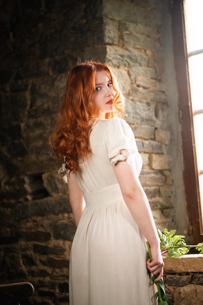 Hermosa chica pelirroja cerca de la ventana vieja con un vestido de noche blanco en una casa tradicional francesa el tema del romance