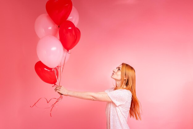 Hermosa chica pelirroja con globo de corazón rojo posando. Concepto de feliz día de San Valentín. Foto de estudio de hermosa chica jengibre bailando sobre fondo rosa.
