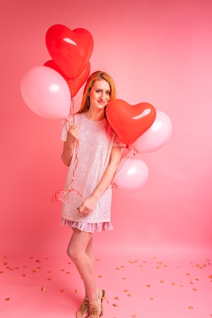 Hermosa chica pelirroja con globo de corazón rojo posando Concepto de feliz día de San Valentín Foto de estudio de hermosa chica jengibre bailando sobre fondo rosa