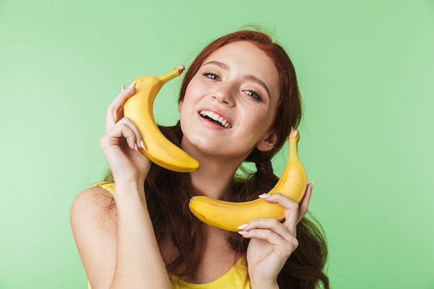 hermosa chica pelirroja joven emocionada posando aislada sobre fondo de pared verde con plátanos frutas imagina hablar de llamada de teléfono móvil.