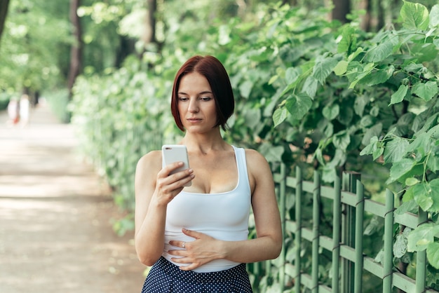 Hermosa chica pelirroja lee pensativamente un mensaje en un teléfono inteligente mientras camina en el parque.
