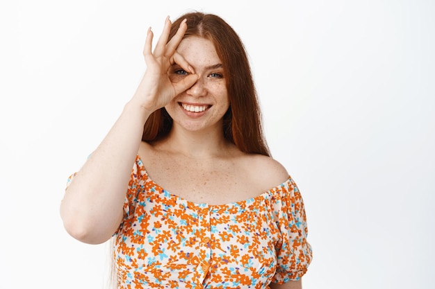 Hermosa chica pelirroja mostrando bien cero OK gesto en el ojo sonriendo complacido recomendando algo bueno fondo blanco