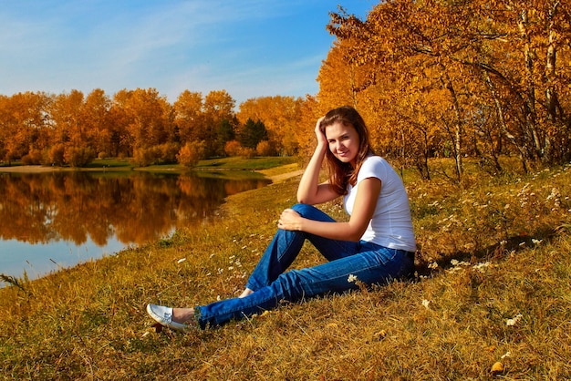 Hermosa chica pelirroja sentada en el suelo en el bosque de otoño junto al lago