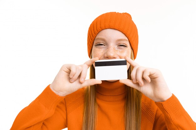 Hermosa chica pelirroja en un sombrero naranja tiene una tarjeta de crédito en un blanco aislado