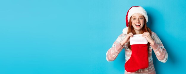 Foto hermosa chica pelirroja con sombrero de santa abre el calcetín de navidad y parece sorprendida de recibir un gif de navidad