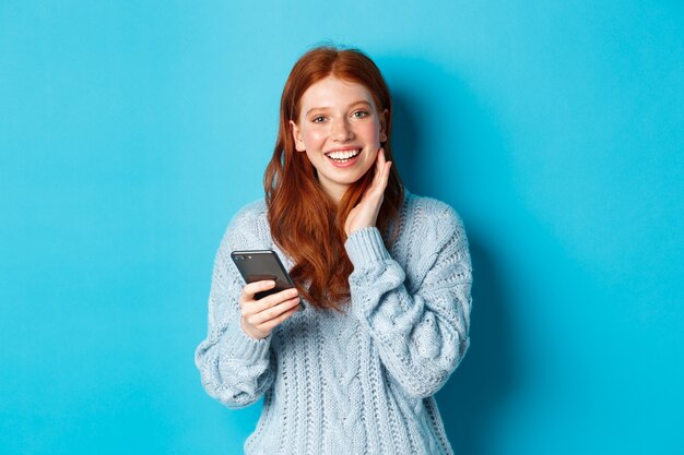 Hermosa chica pelirroja en suéter, sonriendo a la cámara, usando la aplicación de teléfono móvil, de pie con el teléfono inteligente sobre fondo azul.