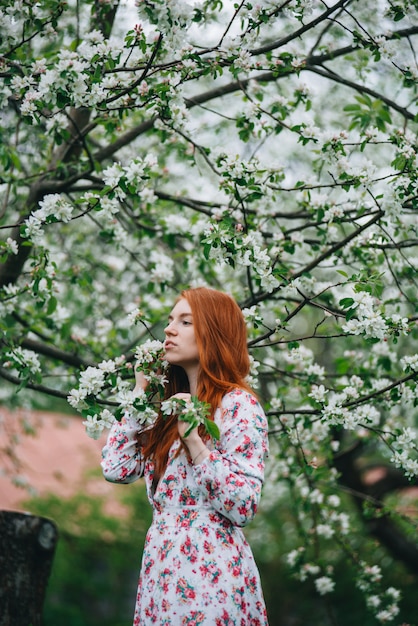 Hermosa chica pelirroja con un vestido blanco entre florecientes manzanos en el jardín.