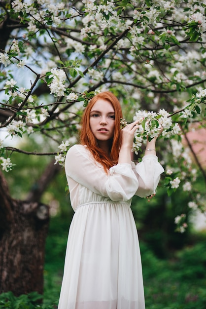 Hermosa chica pelirroja con un vestido blanco entre florecientes manzanos en el jardín