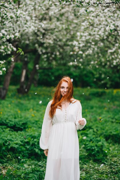 Hermosa chica pelirroja con un vestido blanco entre florecientes manzanos en el jardín