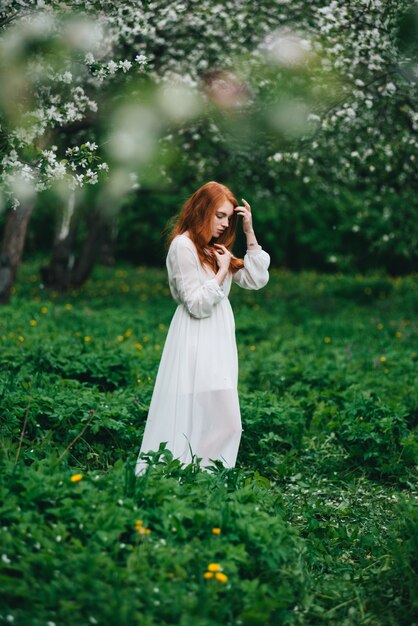 Hermosa chica pelirroja con un vestido blanco entre florecientes manzanos en el jardín