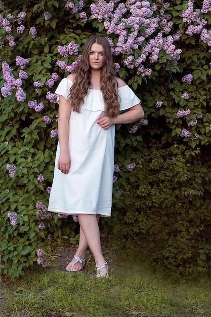 una hermosa chica de pelo largo y castaño con un vestido blanco se encuentra junto a un arbusto lila