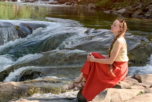 Hermosa chica de pelo largo en falda roja sentada en una roca en una cascada de río de montaña