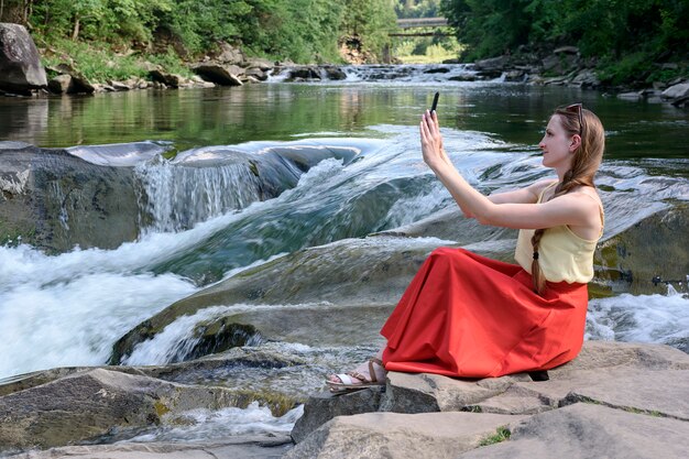 Hermosa chica de pelo largo en una falda roja sentada en una roca y haciendo selfie en el espacio de una cascada del río de montaña. Tarde de verano