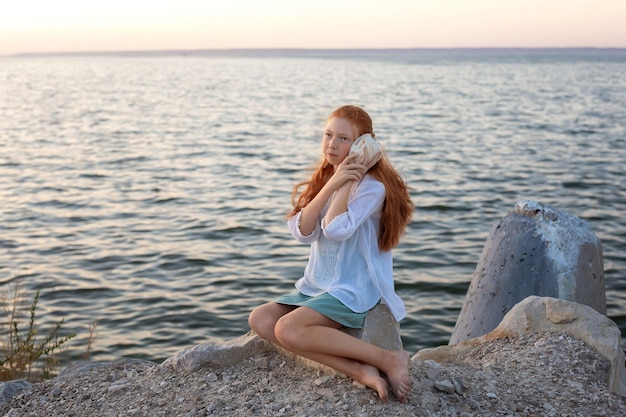 Una hermosa chica con el pelo largo y rojo se sienta en la orilla del río del océano y escucha el sonido del mar desde una gran concha