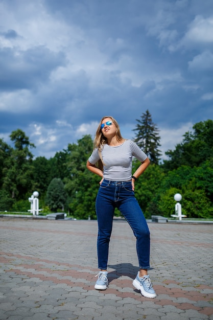 Una hermosa chica de pelo largo rubio de apariencia europea. Vestida con jeans y una camiseta. Caminar en un día soleado