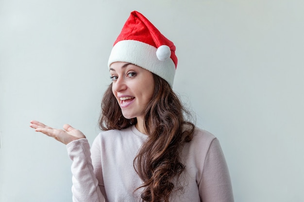 Hermosa chica con el pelo largo en el sombrero rojo de Santa Claus mostrando la palma de la mano abierta aislada sobre fondo blanco. Retrato de mujer joven, verdaderas emociones. Felices vacaciones de Navidad y año nuevo.