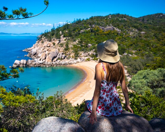 hermosa chica de pelo largo con vestido y sombrero se sienta en la cima de la montaña con vistas a la playa del paraíso