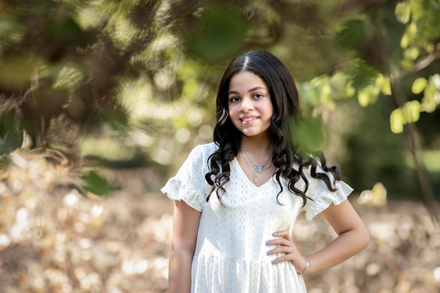 Hermosa chica de pelo negro de pie en la naturaleza y con un vestido blanco, mirando a la cámara