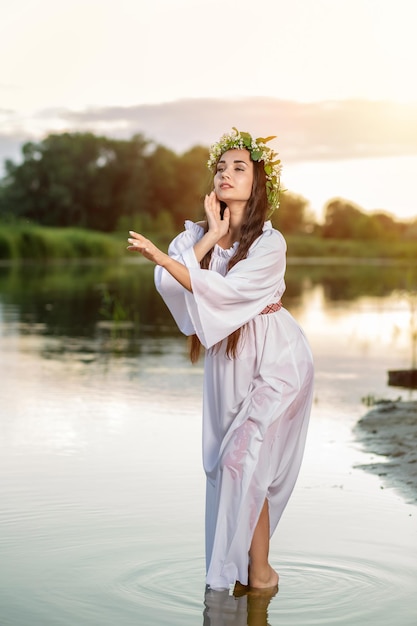 Hermosa chica de pelo negro con vestido blanco vintage y corona de flores en el agua del lago. Llamarada del sol, destello solar.